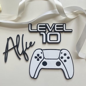 May include: A cake topper set with a gaming theme, featuring the name "Alfie" in black script, the words "LEVEL 10" in a bold font, and a white game controller.