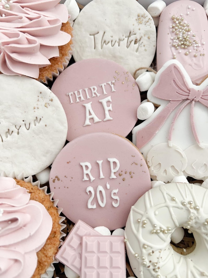 May include: A pink and white birthday gift box with cupcakes, cookies, and a donut. The cookies have the text "Thirty AF" and "RIP 20's" on them.