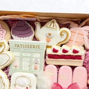 May include: A box filled with decorated sugar cookies, including a teapot, a cupcake, a pastry shop, a strawberry shortcake, and a donut. The cookies are decorated with pink, white, and gold icing.