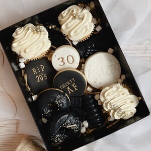 May include: A black gift box filled with black and white cookies and cupcakes. The cookies have gold lettering that says "RIP 20's", "30", "Thirty AF", and "Frazer". The cupcakes have white frosting with gold sprinkles.