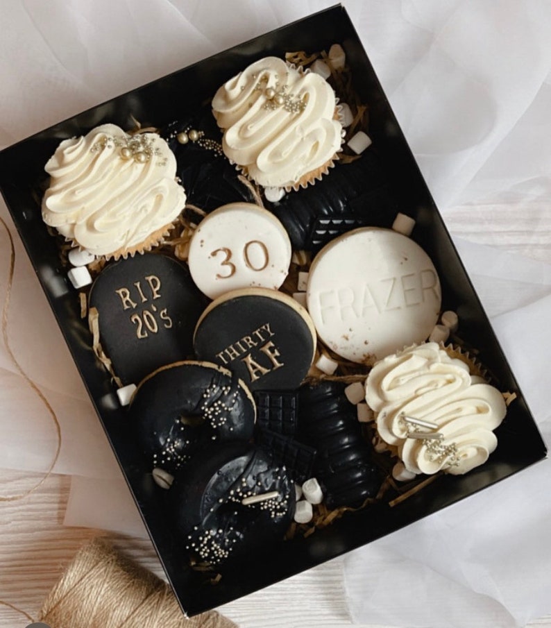 May include: A black gift box filled with black and white cookies and cupcakes. The cookies have gold lettering that says "RIP 20's", "30", "Thirty AF", and "Frazer". The cupcakes have white frosting with gold sprinkles.