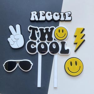 May include: Cake toppers in black and white. The toppers feature the name "Reggie", the phrase "Two Cool", a peace sign, sunglasses, a lightning bolt, and two smiley faces.