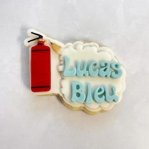 May include: A white cookie decorated with red and gold icing depicting a fire extinguisher. The cookie also has blue icing with the text "Lucas Bleu" on it.