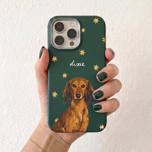 Könnte beinhalten: Eine dunkelgrüne Handyhülle mit einer gemalten Illustration eines langhaarigen braunen Dackels. Über dem Hund steht der Name "dixie" in weißer Schreibschrift, und die Hülle ist mit kleinen, verstreuten goldfarbenen Sternen verziert.