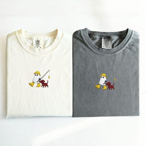 May include: Two folded t-shirts, one cream and one gray, featuring an embroidered design. The design depicts a ghost-like figure in a yellow hat and boots fishing with a small brown dog. The shirts have the "Comfort Colors" logo.