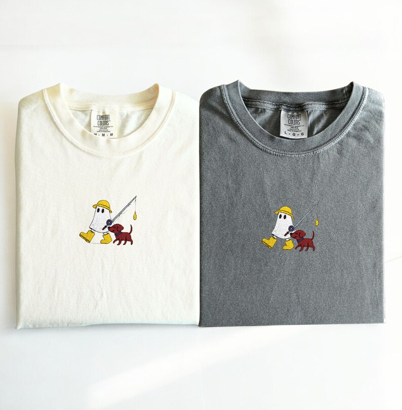 May include: Two folded t-shirts, one cream and one gray, featuring an embroidered design. The design depicts a ghost-like figure in a yellow hat and boots fishing with a small brown dog. The shirts have the "Comfort Colors" logo.