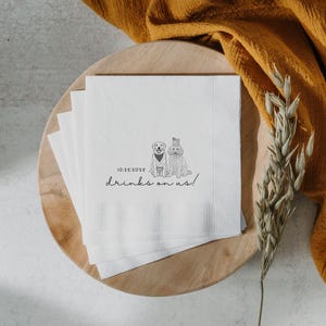 May include: White paper napkins with a black illustration of two dogs, one wearing a bow tie and the other a hat. The text "10.25.2025 drinks on us!" is printed below the dogs. The napkins are on a wooden plate.