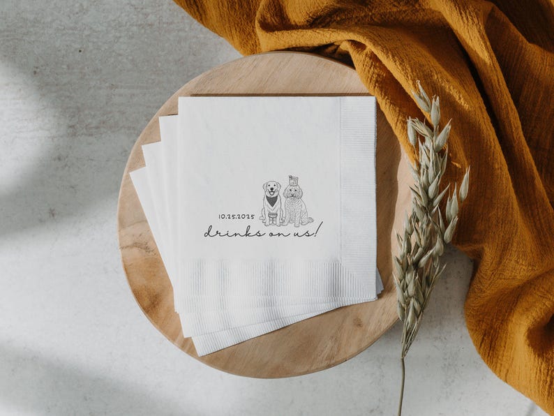 May include: White paper napkins with a black illustration of two dogs, one wearing a bow tie and the other a hat. The text "10.25.2025 drinks on us!" is printed below the dogs. The napkins are on a wooden plate.