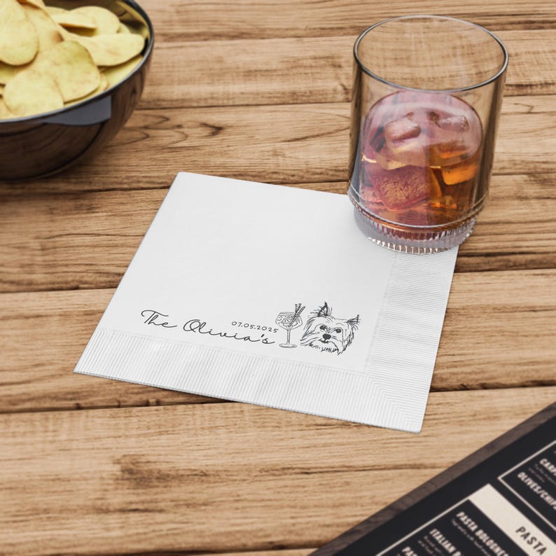 May include: A white cocktail napkin with a decorative border, featuring the text "The Olivia's" and the date "07.05.2025". A sketched illustration of a cocktail and a dog is also present. A glass of amber liquid with ice sits nearby.