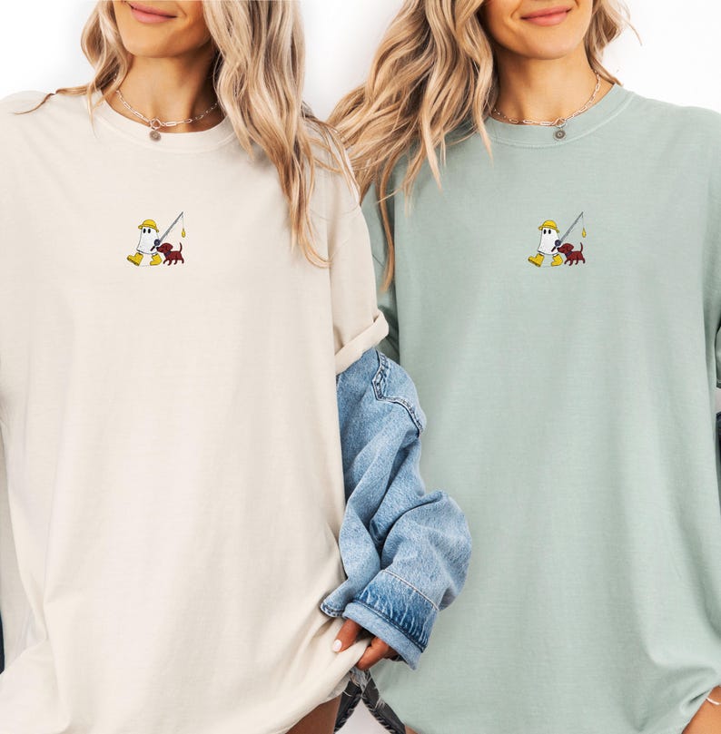 May include: Two long-sleeve t-shirts, one cream and one sage green, each featuring an embroidered cartoon design of a figure fishing with a dog. The figure wears a yellow hat and boots.