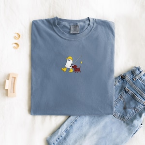 May include: A blue t-shirt featuring an embroidered design of a ghost wearing a yellow hard hat and boots, fishing with a red dachshund. The shirt is folded with a pair of jeans and accessories in the background.