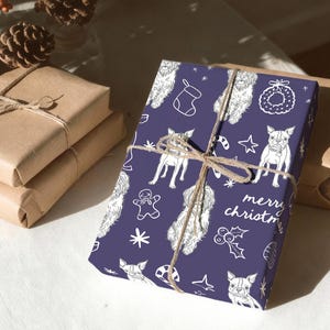May include: A rectangular gift wrapped in dark purple paper featuring a white line art pattern of dogs, gingerbread men, stockings, wreaths, and holly, tied with brown twine. The text "merry christmas" is printed in white on the paper.
