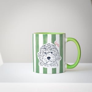 May include: A ceramic mug with a green and white striped pattern. The mug features a line drawing of a dog's face and the name "Lola" in pink. The handle and interior of the mug are green.