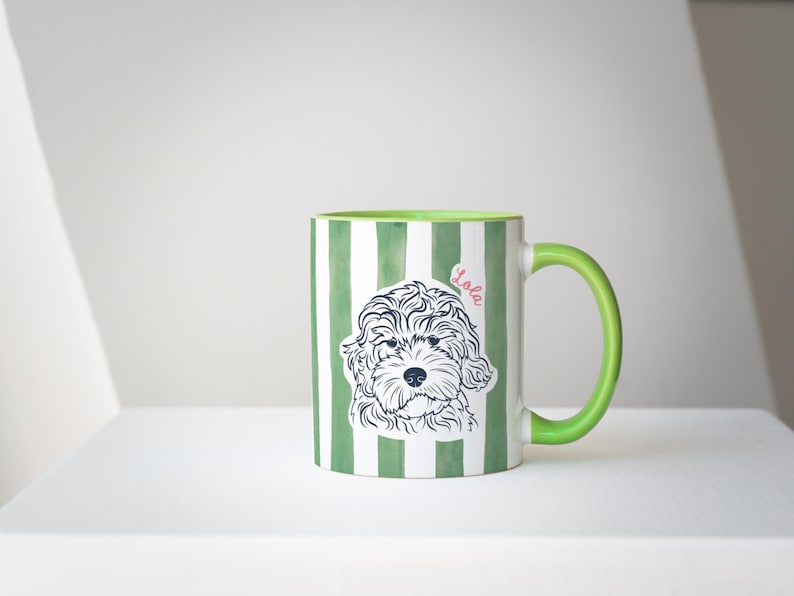 May include: A ceramic mug with a green and white striped pattern. The mug features a line drawing of a dog's face and the name "Lola" in pink. The handle and interior of the mug are green.
