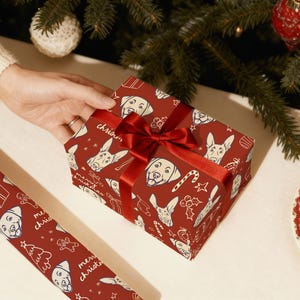 May include: A rectangular gift box wrapped in deep red paper featuring a repeating white line-art pattern of dog faces, candy canes, and the text merry christmas, tied with a shiny red satin ribbon bow, alongside a matching roll of wrapping paper.