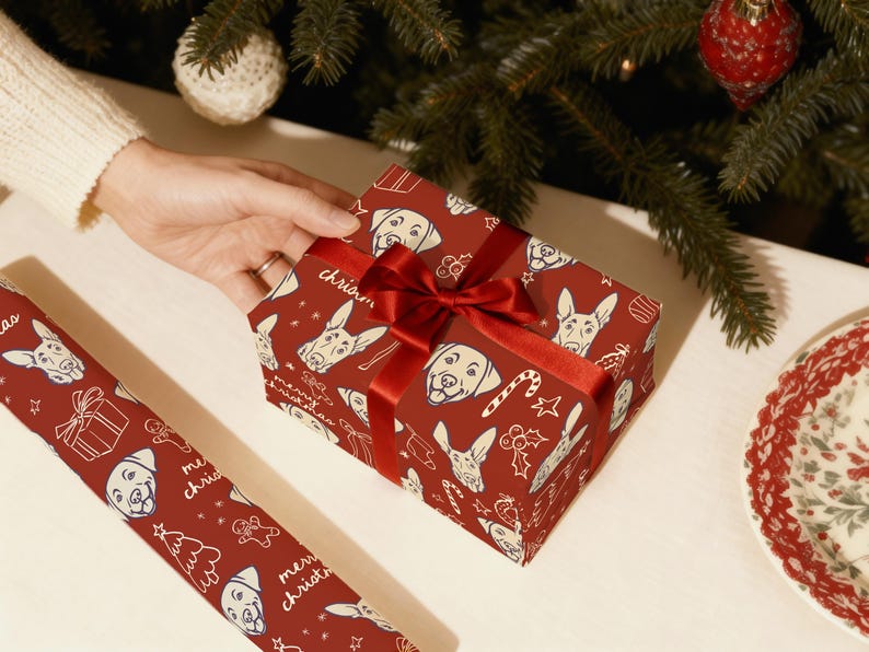 May include: A rectangular gift box wrapped in deep red paper featuring a repeating white line-art pattern of dog faces, candy canes, and the text merry christmas, tied with a shiny red satin ribbon bow, alongside a matching roll of wrapping paper.