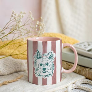 May include: A ceramic mug with a pink interior and handle, featuring vertical white and pink stripes. The mug has a sketch of a dog with the word "Ohana" above it. The mug is on a white knitted surface.