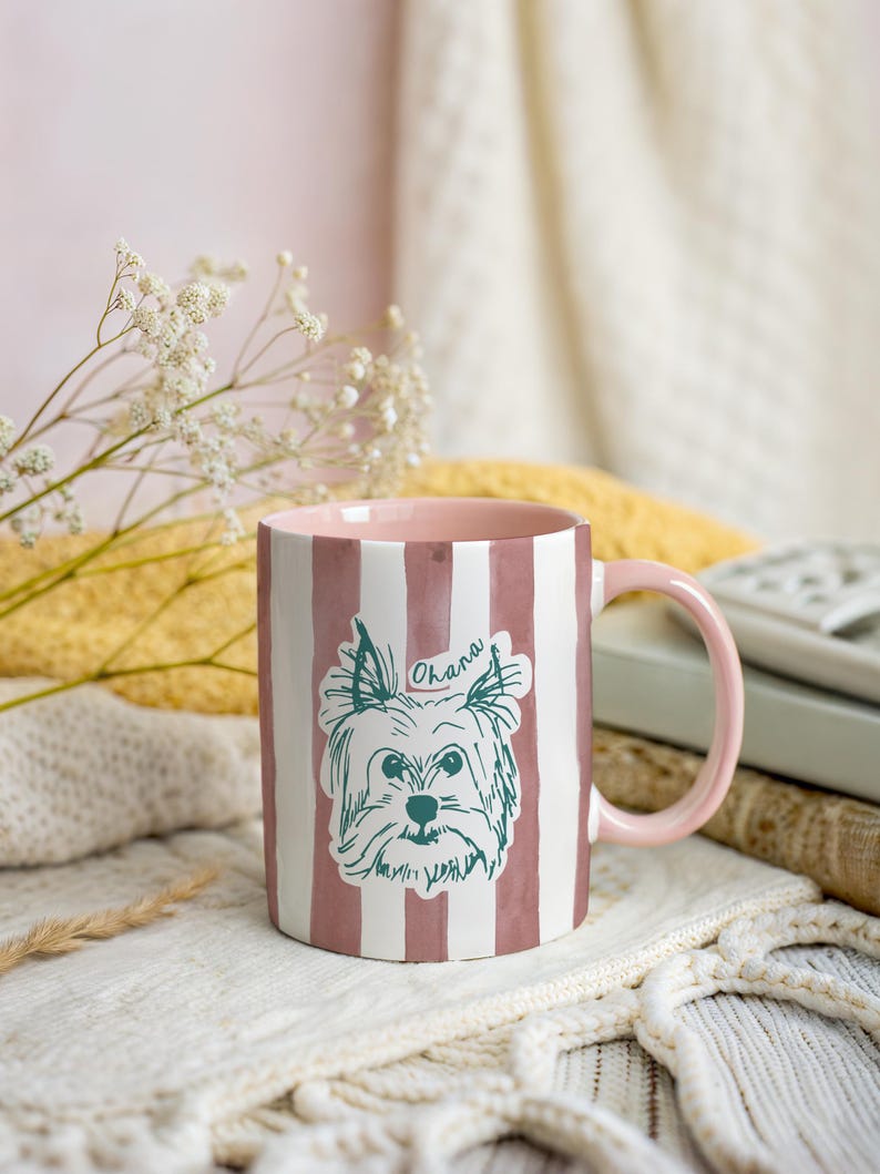 May include: A ceramic mug with a pink interior and handle, featuring vertical white and pink stripes. The mug has a sketch of a dog with the word "Ohana" above it. The mug is on a white knitted surface.