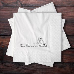 May include: White paper napkins with a black illustration of a Basset Hound and the text "The Oliver's" and the date "07.05.2025". The napkins are stacked on a dark wood surface.