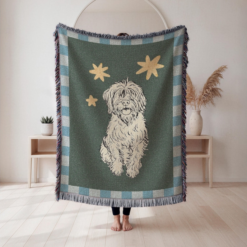 May include: A woven throw blanket featuring a cream-colored dog illustration with three gold stars on a dark teal background. The blanket has a blue and white checkered border with fringe.