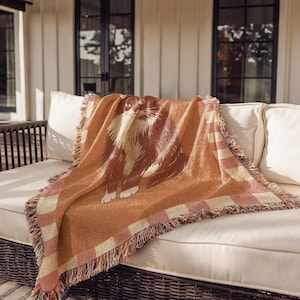 May include: A woven throw blanket featuring a large cat illustration in shades of brown and cream. The blanket has a fringed edge and a checkered border in pink and cream. The throw is draped over a white outdoor sofa.