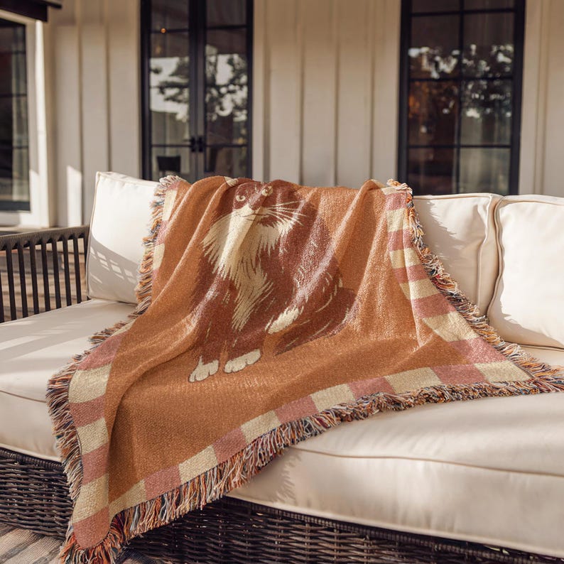 May include: A woven throw blanket featuring a large cat illustration in shades of brown and cream. The blanket has a fringed edge and a checkered border in pink and cream. The throw is draped over a white outdoor sofa.