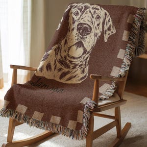 May include: A brown throw blanket draped over a wooden rocking chair. The blanket features a large dog portrait in beige and brown, with the name "Samson" in script. The blanket has a checkered border and fringed edges.