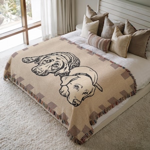 May include: Beige throw blanket featuring two dog portraits in black line art. The blanket has a geometric border and fringe trim. The throw is displayed on a bed with pillows.