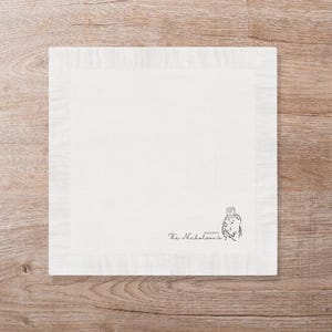 May include: A square, off-white linen napkin with a fringed border. The lower right corner features a small illustration and the text "The Nicholson's". The napkin is set on a wooden surface.