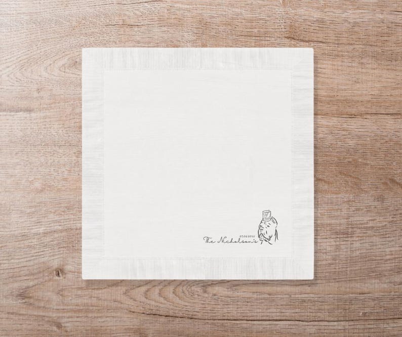 May include: A square, off-white linen napkin with a fringed border. The lower right corner features a small illustration and the text "The Nicholson's". The napkin is set on a wooden surface.