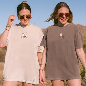 May include: Two women wearing oversized t-shirts. One shirt is beige, the other is brown. Both shirts feature a small embroidered design of a cartoon character with a fishing rod and a dog. The background is a desert landscape.