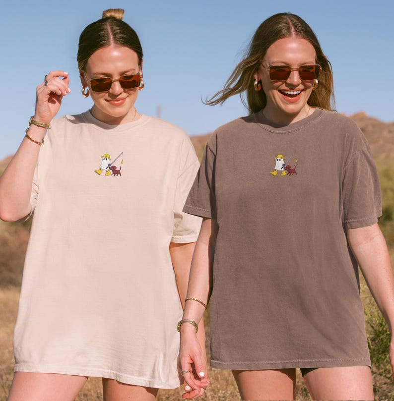 May include: Two women wearing oversized t-shirts. One shirt is beige, the other is brown. Both shirts feature a small embroidered design of a cartoon character with a fishing rod and a dog. The background is a desert landscape.