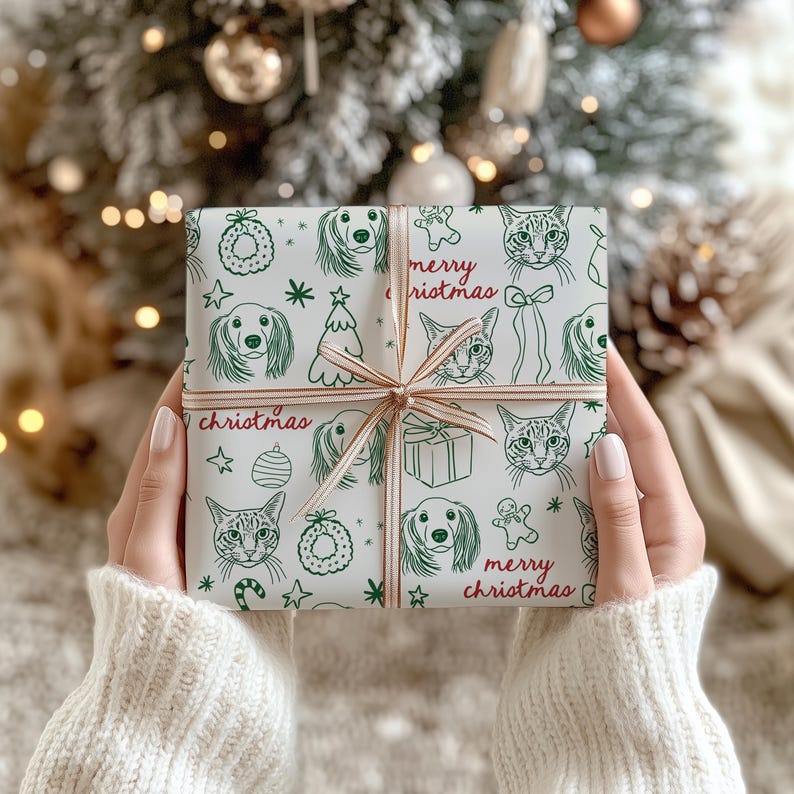 May include: A square gift box wrapped in white paper featuring a repeating green line-art pattern of dogs, cats, Christmas trees, wreaths, and gingerbread men, with the text merry christmas in red, tied with a thin, light-colored ribbon.