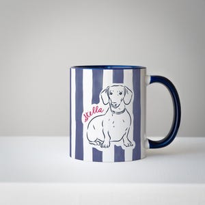 May include: A ceramic mug with a blue and white striped pattern. The mug features a line drawing of a dachshund and the name "Stella" in pink script. The handle and interior rim are navy blue.