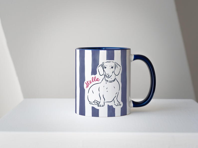May include: A ceramic mug with a blue and white striped pattern. The mug features a line drawing of a dachshund and the name "Stella" in pink script. The handle and interior rim are navy blue.