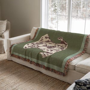 May include: A green throw blanket with a cream-colored abstract design draped over a white sofa. The blanket has a fringed edge with brown, orange, and cream accents. The sofa is in a room with a window overlooking a snowy landscape.