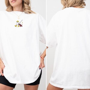 May include: White oversized t-shirt featuring a small embroidered design of a figure fishing with a dog. The design is in yellow, brown, and red. The shirt has short sleeves and a relaxed fit, suitable for casual wear.