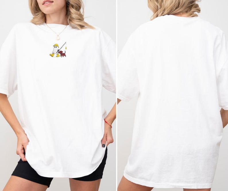 May include: White oversized t-shirt featuring a small embroidered design of a figure fishing with a dog. The design is in yellow, brown, and red. The shirt has short sleeves and a relaxed fit, suitable for casual wear.