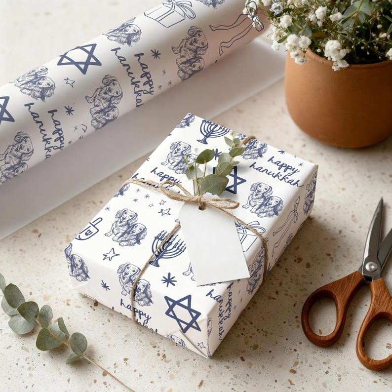 May include: White wrapping paper featuring a repeating dark blue pattern of dachshund dogs, menorahs, Stars of David, and the text "happy hanukkah". A rectangular gift is wrapped in this paper, tied with twine, and includes a blank white gift tag.