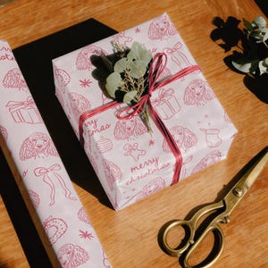May include: A roll of light pink wrapping paper and a square gift box wrapped in the same paper, both featuring a repeating red line-art pattern of poodles, gift boxes, and the text merry christmas, tied with red twine and topped with green leaves.