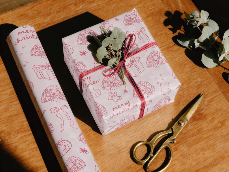 May include: A roll of light pink wrapping paper and a square gift box wrapped in the same paper, both featuring a repeating red line-art pattern of poodles, gift boxes, and the text merry christmas, tied with red twine and topped with green leaves.