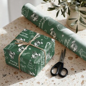 May include: A square gift box wrapped in dark green paper with a white line-art pattern of dog faces, Christmas trees, candy canes, and the text "merry christmas," tied with brown twine, next to a matching roll of paper and black scissors.