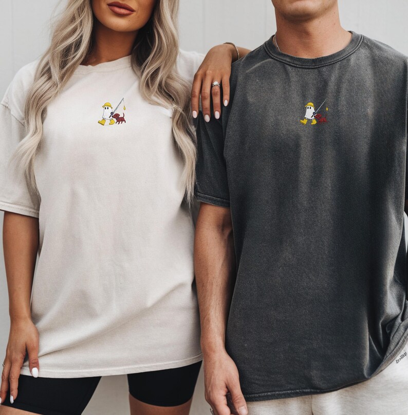 May include: Two short-sleeved t-shirts, one cream and one charcoal, featuring a small embroidered design of a cartoon character fishing. The character wears a yellow hat and boots. The shirts are a relaxed fit.