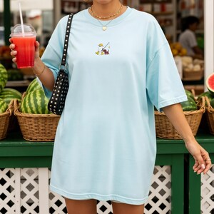 May include: Light blue t-shirt with a small embroidered design of a cartoon character fishing with a crab. The shirt is a relaxed fit. The person is holding a drink in a clear plastic cup with a red liquid and a straw. A black studded purse hangs from the shoulder.