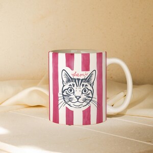 May include: White ceramic mug with a pink striped pattern and a black and white illustration of a cat's face. The name "Sami" is written above the cat's head in cursive.