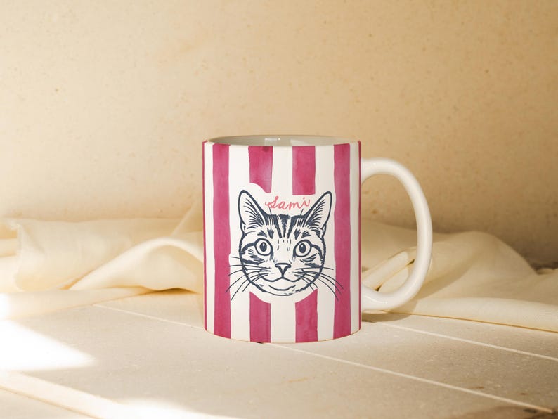 May include: White ceramic mug with a pink striped pattern and a black and white illustration of a cat's face. The name "Sami" is written above the cat's head in cursive.