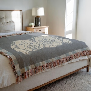 May include: A gray woven throw blanket featuring a cream-colored illustration of two dogs. The blanket has a fringed edge with a plaid border in shades of blue, red, and cream. The blanket is draped over a bed.