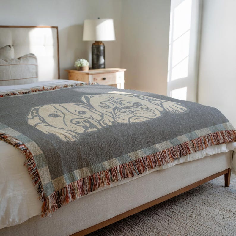 May include: A gray woven throw blanket featuring a cream-colored illustration of two dogs. The blanket has a fringed edge with a plaid border in shades of blue, red, and cream. The blanket is draped over a bed.