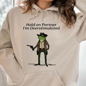 May include: Beige hoodie with a cartoon frog cowboy graphic. The frog is green with a brown hat, jacket, and green trousers, holding a gun. Text above the frog reads "Hold on Partner I'm Overstimulated."