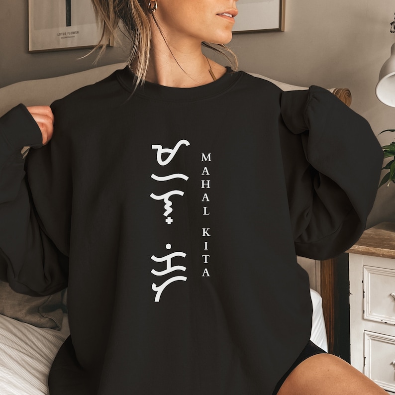 May include: A black crewneck sweatshirt featuring white text printed vertically. The design includes Baybayin script characters on the left and the words MAHAL KITA written in a vertical column on the right side of the chest area.